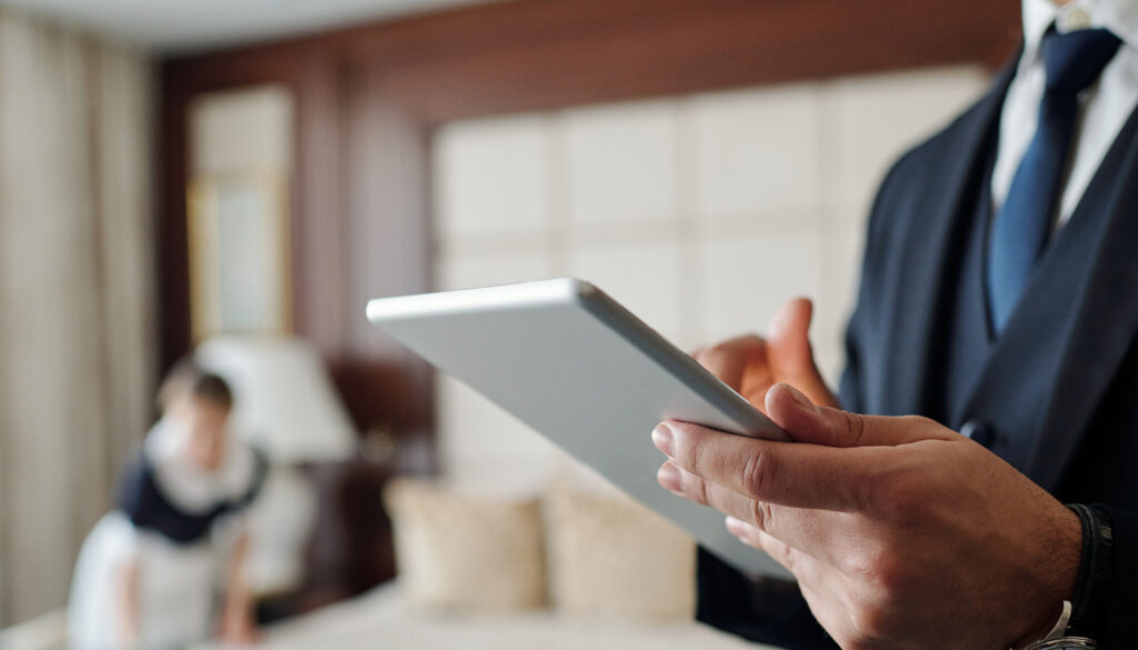 Profissional de hotelaria usando tablet em quarto de hotel, avaliando checklist de enxoval para alta temporada de verão