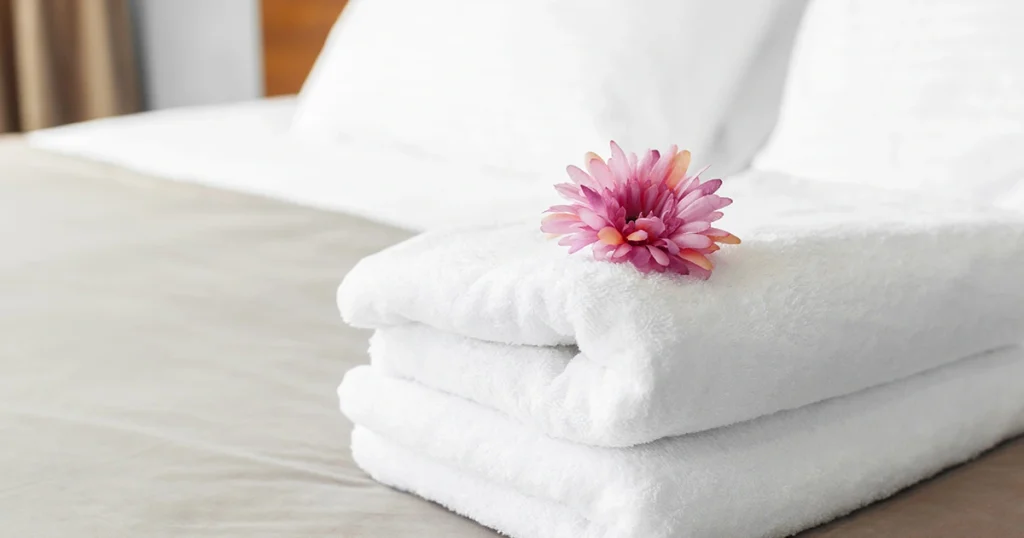 Toalhas brancas para hotelaria sobre cama com flor decorativa em destaque