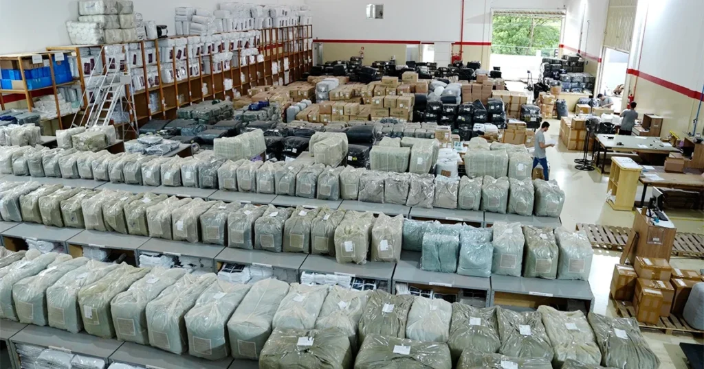 Vista ampla de um centro de distribuição com centenas de pacotes e produtos organizados em prateleiras e mesas, enquanto funcionários realizam a separação e logística dos pedidos.