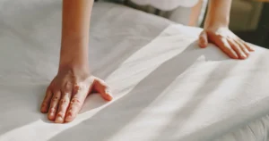 Mãos ajustando lençol branco em cama de hotel com enxoval profissional para hotelaria premium
