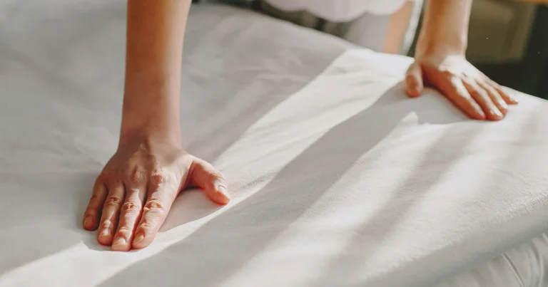 Mãos ajustando lençol branco em cama de hotel com enxoval profissional para hotelaria premium